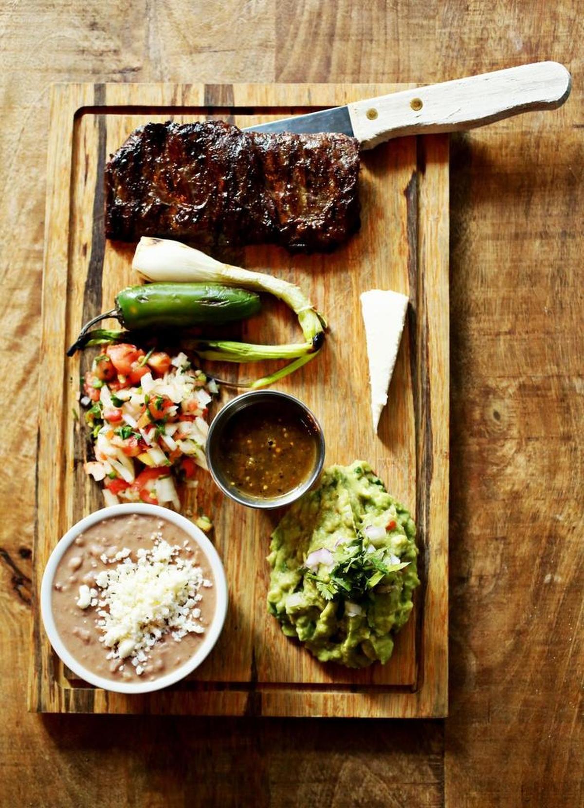 The Una Asada entree will score you an ample slab of carne asada (grilled skirt steak), a wedge of queso fresco and a small bowl of casa beans (aka refried beans), laid out on a carving board garnished with a grilled jalapeño, grilled scallion and mounds of guacamole and pico de gallo at La Santa in Raleigh.