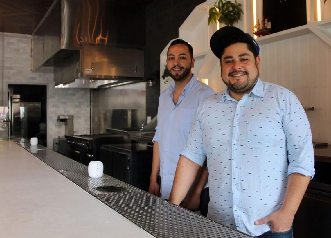 A welcoming portrait of two chefs at their restaurant, Cortez. A smiling chef in a patterned shirt and backwards baseball cap is in the foreground, while a second one stands behind him. They are positioned behind a modern, light-colored bar, with the restaurant’s professional open kitchen visible in the background.