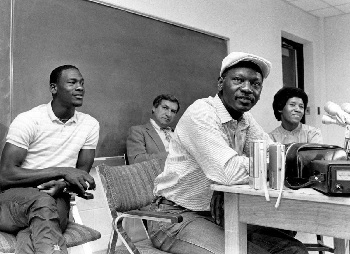 Michael Jordan (left) looks on as his father James Jordan speaks at a press conference May 5, 1984 where Jordan announced that he was entering the NBA draft. At rear is UNC coach Dean Smith and at right is Jordan’s mother Delores.