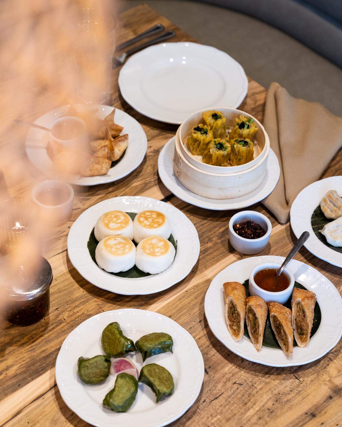 A high-angle shot of a wooden table covered with a variety of dim sum dishes. In the center, a bamboo steamer holds yellow siu mai, surrounded by white plates featuring pan-fried buns, fried spring rolls cut in half, green crescent dumplings, and fried wontons. Several small dipping sauce bowls, including one with chili oil, are also on the table.