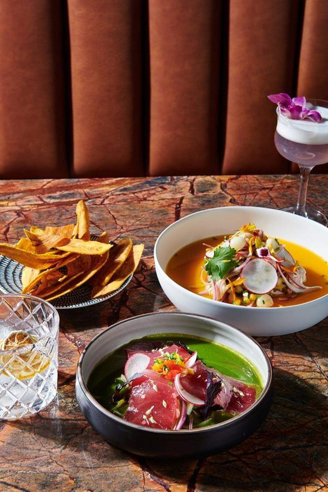 A moody, professional shot of a meal on a red-veined marble table. The dishes include a bowl of raw tuna in a green leche de tigre and another bowl of ceviche. The meal is accompanied by plantain chips and two craft cocktails, one of which is a purple, frothy drink garnished with an orchid.