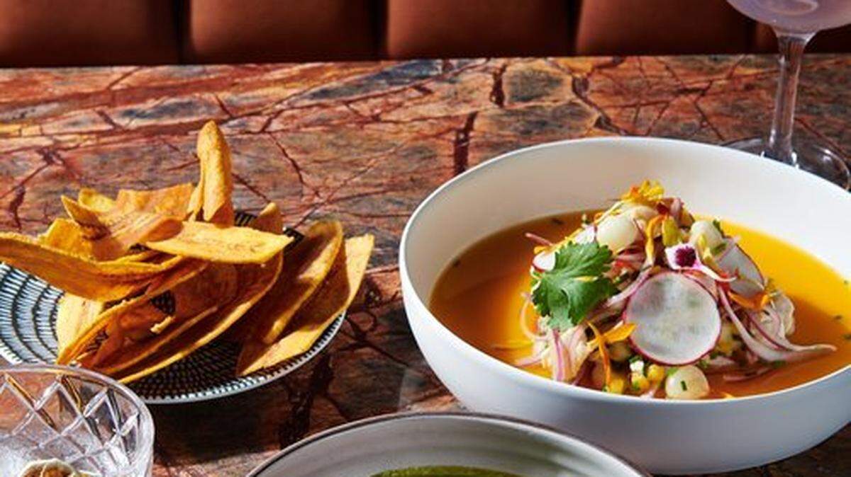 A moody, professional shot of a meal on a red-veined marble table. The dishes include a bowl of raw tuna in a green leche de tigre and another bowl of ceviche. The meal is accompanied by plantain chips and two craft cocktails, one of which is a purple, frothy drink garnished with an orchid.