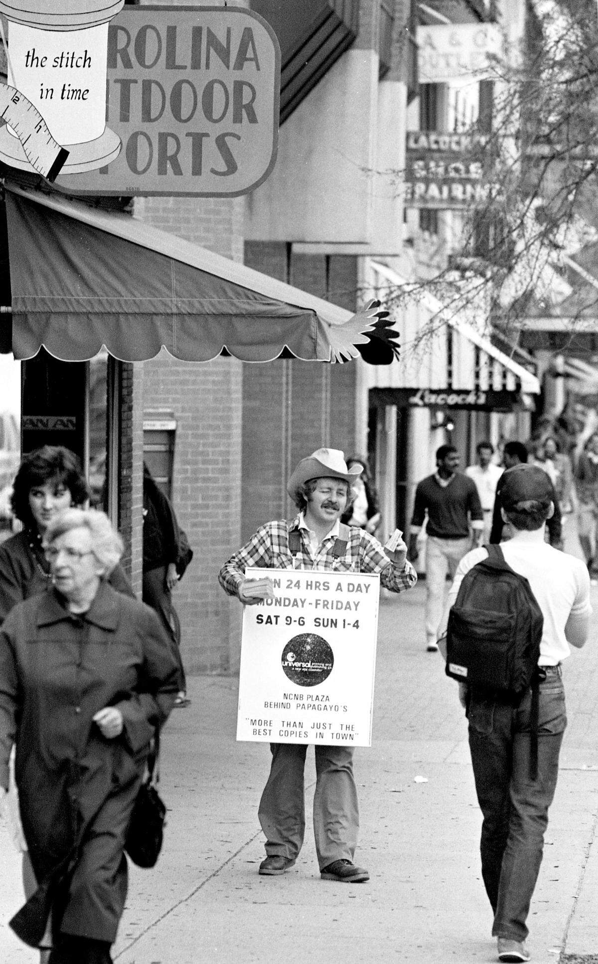 Image of a 1980’s era street vendor with sandwich board advertising and promoting the services of Universal Printing in the NCNB Plaza on Franklin Street in Chapel Hill, N.C. From the News & Observer archives