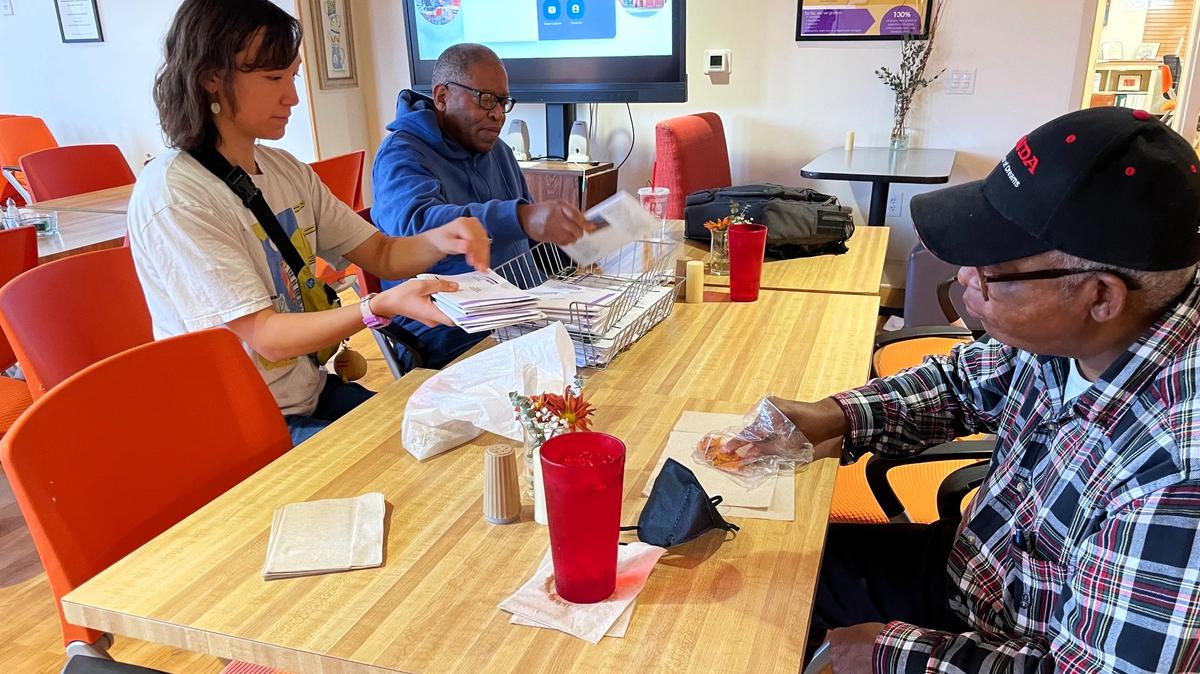 A Club Nova colleague helps staff member M. Ren-Wen Huang prepare copies of the monthly Stargazer Gazette newsletter to go in the mail, while another club colleague grabs a quick bite to eat on Monday, Nov. 11, 2024, in Carrboro.