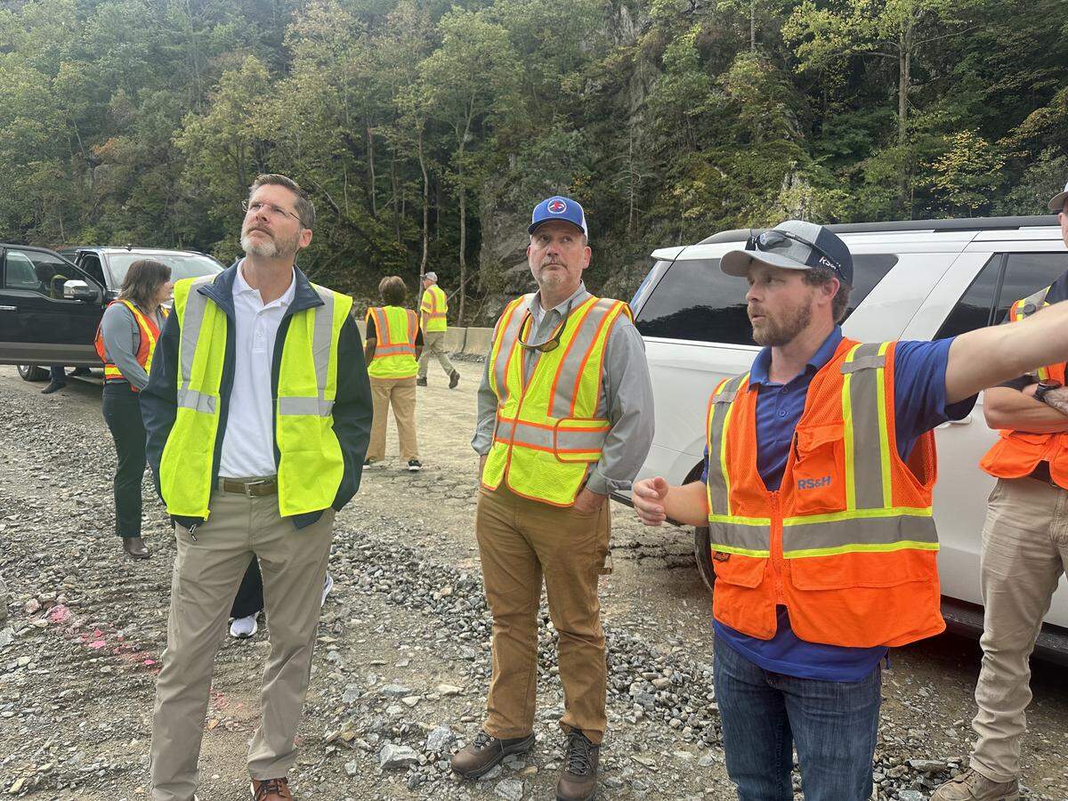 Federal Highway Administrator Sean McMaster, left, and state Transportation Secretary Joey Hopkins, center, look up toward Interstate 40 as project manager Blake Soblesky describes how contractors will rebuild the eastbound lanes washed away by the Pigeon River. McMaster was making his first visit to the gorge on Friday, the one-year anniversary of Helene’s devastating arrival in Western North Carolina.