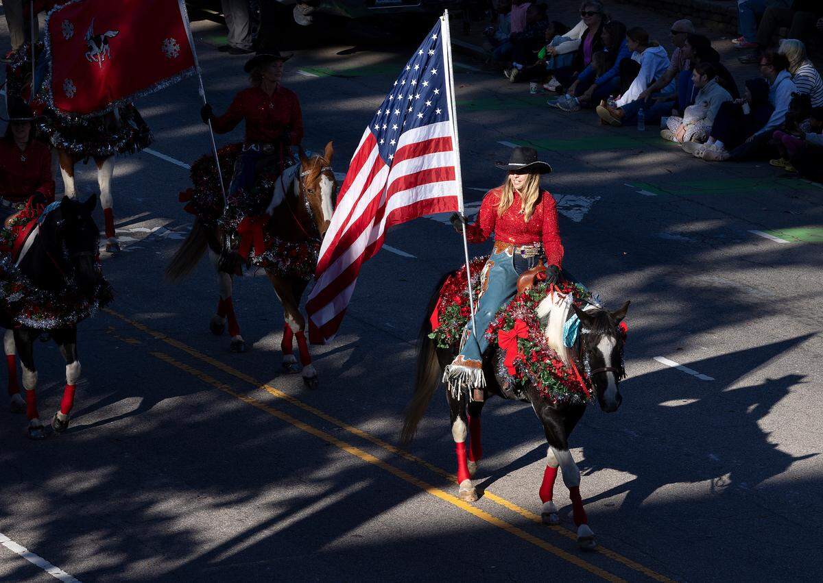 A person carries an American flag while riding horses with Carousel Farms during the Raleigh Christmas Parade on Saturday, Nov. 18, 2023.