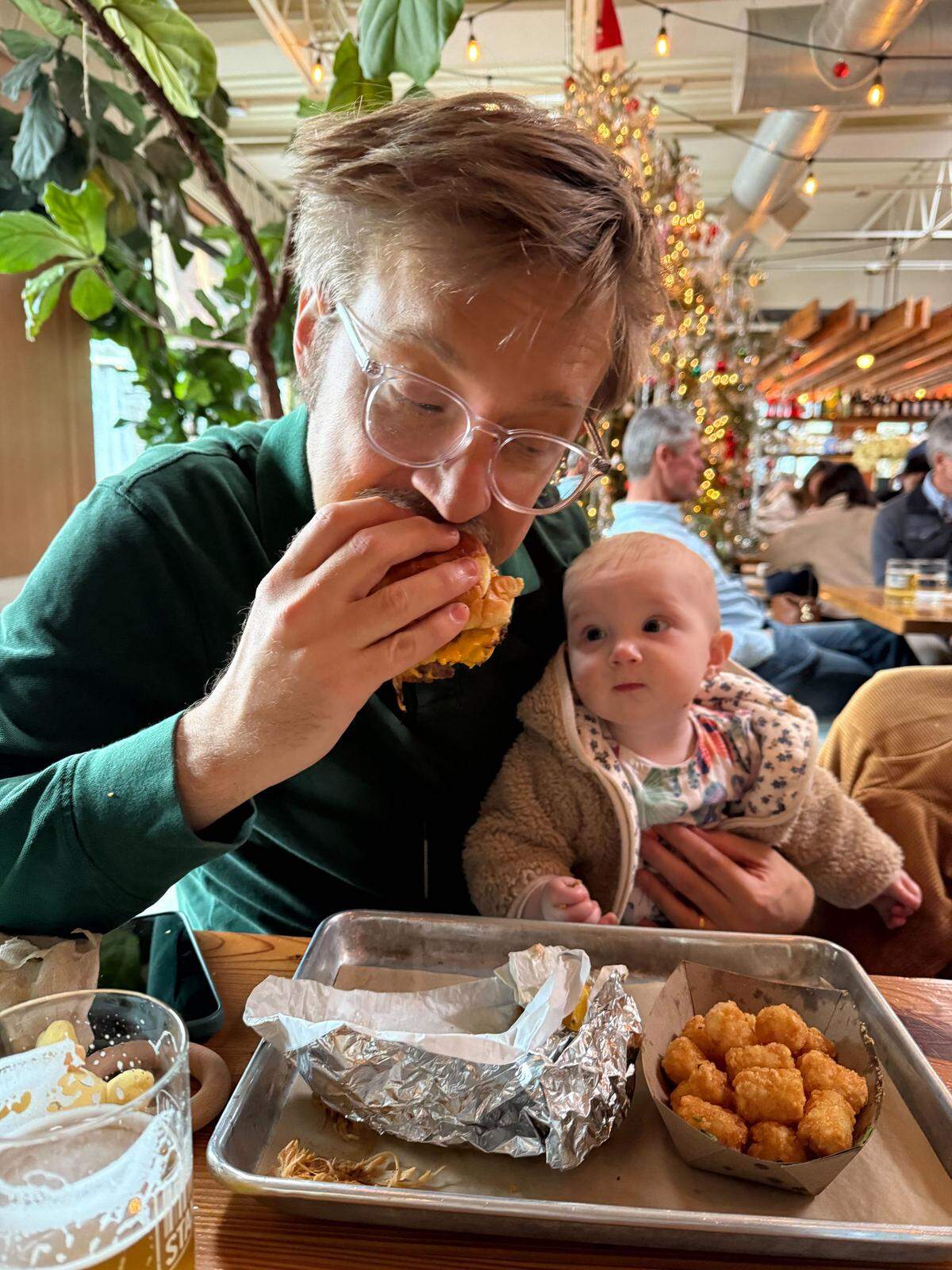 The N&O’s food & dining reporter Drew Jackson, pictured at Standard Beer with his 1-year-old daughter Ella, eating a smashburger and tater tots in December 2025 while reporting the Top 50 Restaurants project. (Photo by Kerry O’Connor, Drew’s wife and willing participant in this months-long journey.)