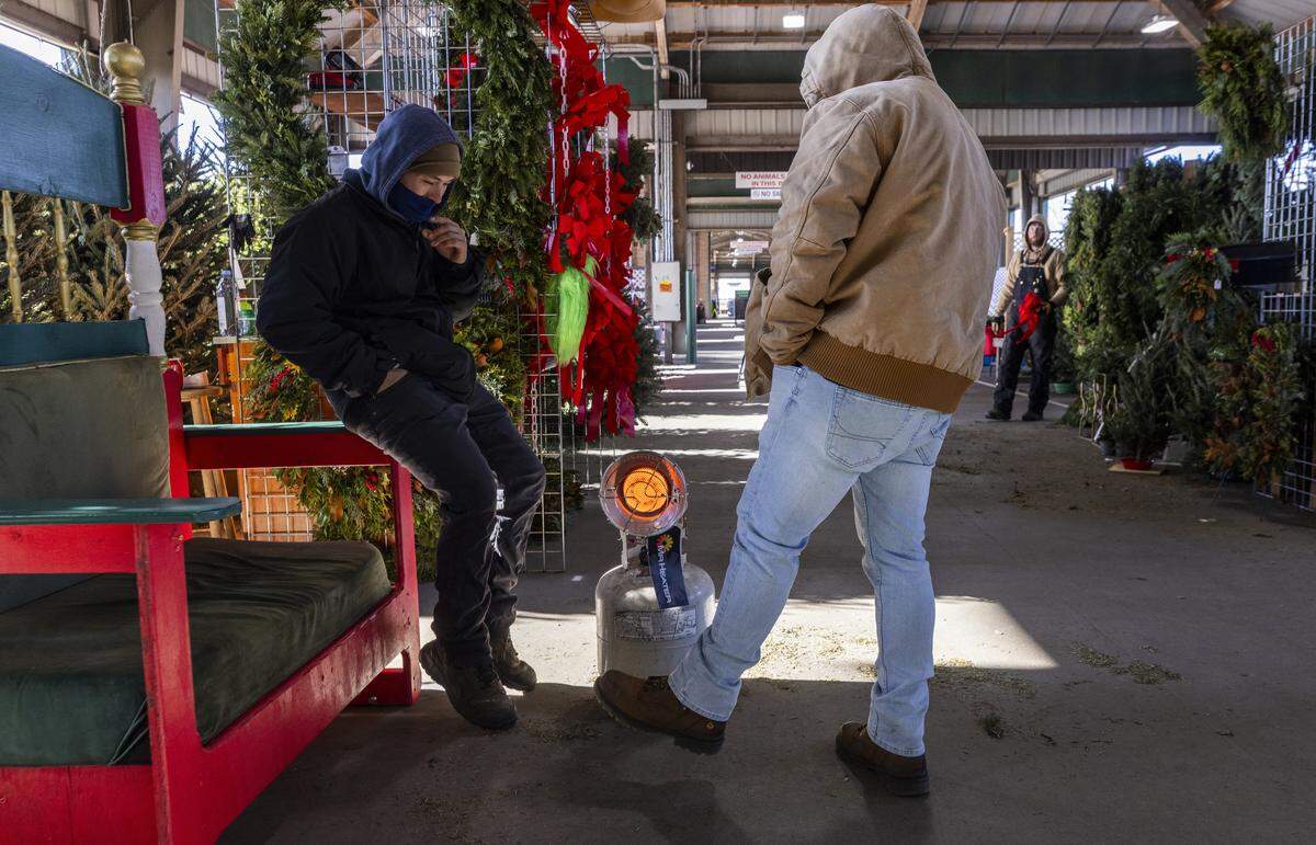 Brothers  Israel, left, and George Delgado of Jernigan’s Nursery in West Jefferson try to keep warm near a propane heater at the company’s Christmas tree vendor stall at the North Carolina State Farmers Market in Raleigh on Monday morning, Dec. 15, 2025. Temperatures in Raleigh dipped into the teens, marking the city’s coldest weather since the winter of 2022.