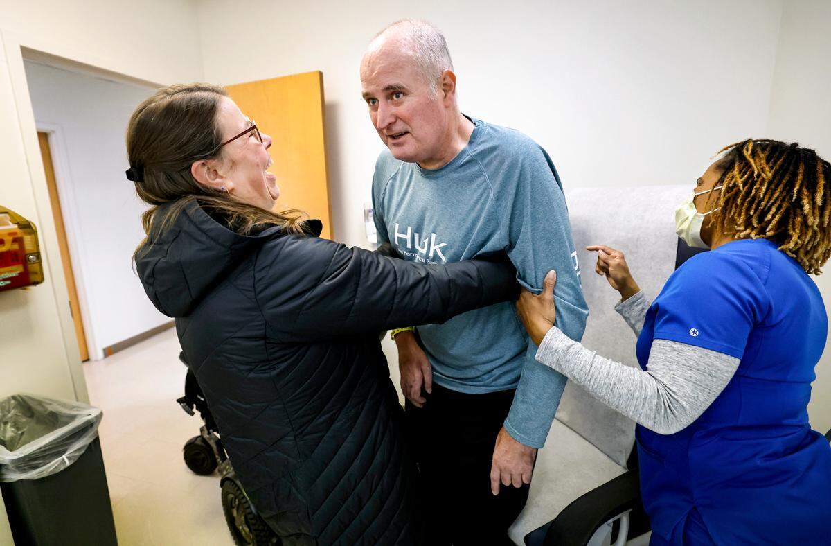 Jamie Woodlief laughs as she helps her husband Roy into the exam room chair during his appointment at the Duke ALS Clinic in Durham, N.C., Tuesday, Jan. 21, 2025. Helping out is Pamela Brown. Roy has been diagnosed with amyotrophic lateral sclerosis (ALS), forcing him to retire from the Wake County Sheriff’s Office. The fatal disease causes muscle weakness, making it difficult to walk, eat and eventually breathe.