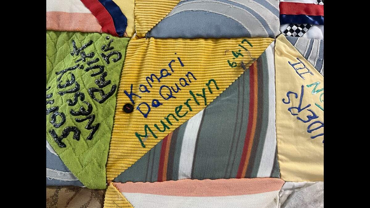 The name “Kamari DaQuan Munerlyn” and the date he was killed written in blue sharpie on one half of a square patch of the Durham Homicide Memorial Quilt. One half is gold with thin stripes. The other has thick vertical stripes of dark teal with thinner stripes of yellow, orange and baby blue.