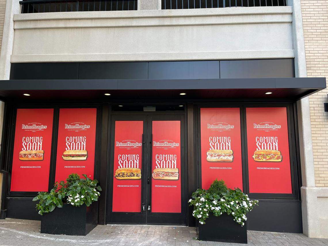 An exterior view of a forthcoming PrimoHoagies location with its windows and double doors covered by bright red promotional banners. The banners feature the white “PrimoHoagies” logo and bold white text reading “COMING SOON” above various images of specialty sandwiches. Two large black planter boxes with white flowers flank the entrance.