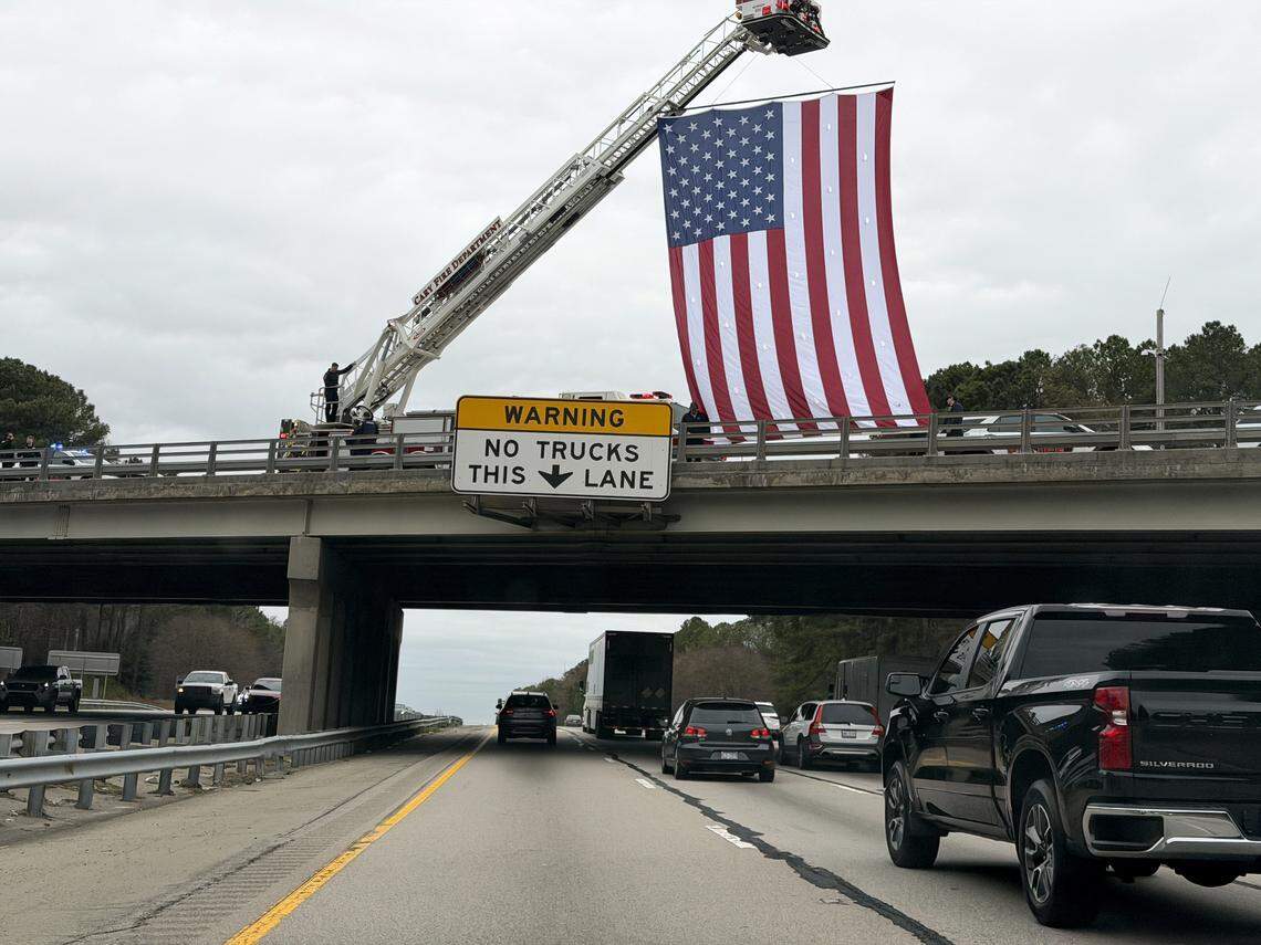 A large U.S. flag is displayed over I-40 as firefighters and law enforcement personnel line an overpass to honor fallen N.C. Highway Patrol Master Trooper Steven Perry as his body was transported with a law enforcement escort on Monday afternoon from the State Medical Examiner's Office in Raleigh to Fisher Funeral Parlor in Durham.