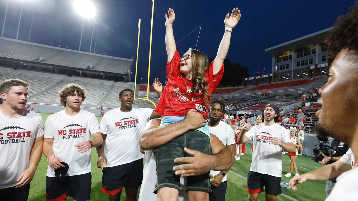 As N.C. State’s Joe Shimko, far left, watches, offensive lineman Rylan Vann lifts up Gabi Angelini of Raleigh after she scored a touchdown during “Victory Day” at Carter-Finley Stadium in Raleigh, N.C., Friday, August 5, 2022. On Victory Day, the Wolfpack partner with GiGi’s Playhouse, giving cognitively and physically impaired children a chance to play football alongside the Pack.
