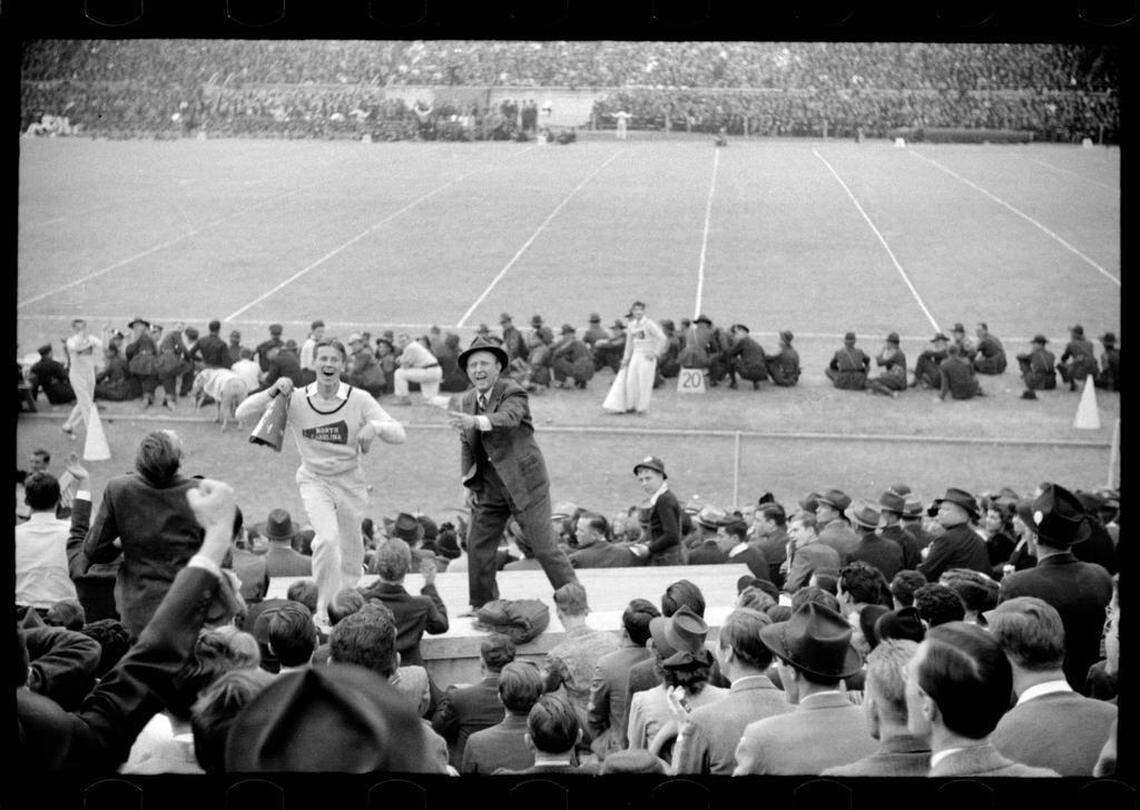 Fans ringed the sidelines and famous bandleader Kay Kyser (right) and a UNC cheerleader lead the crowd in cheers at the 1939 Duke-Carolina game played in Durham, NC. UNC’s mascot “Rameses” can be seen just behind the cheerleader.