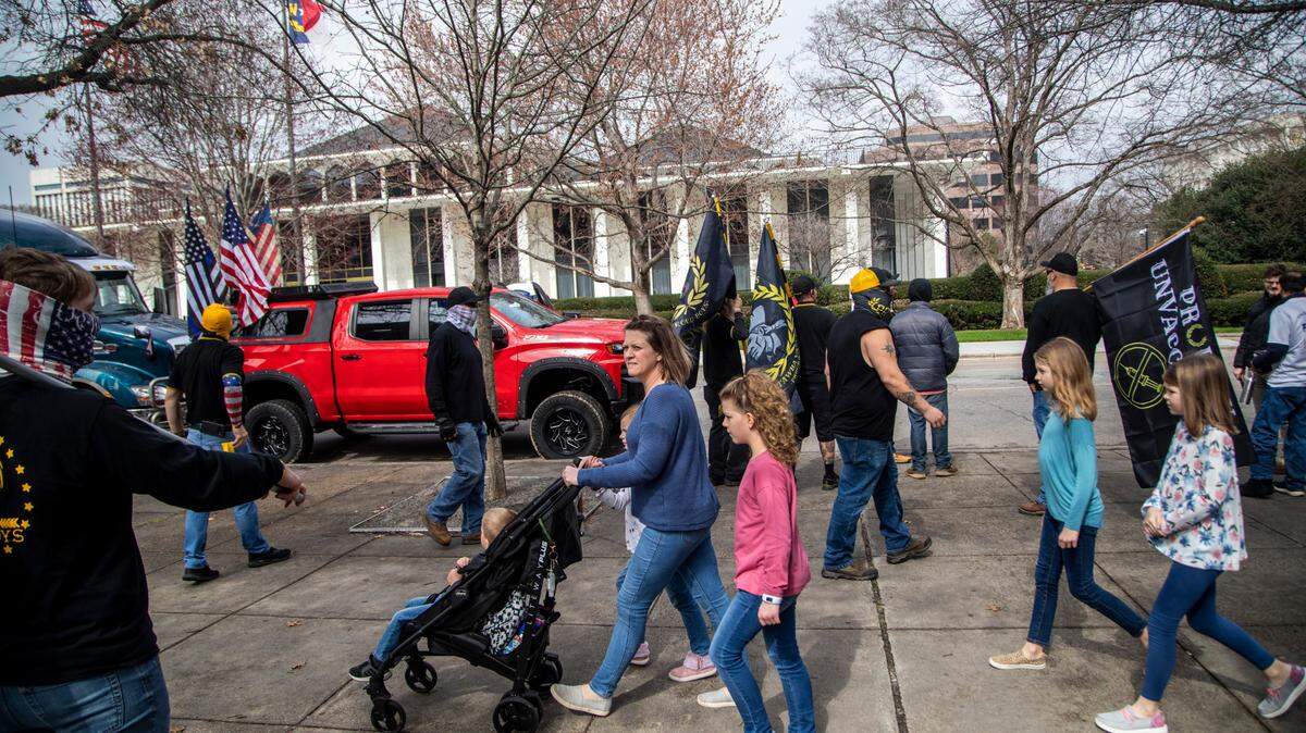 A woman walks with a baby stroller and children past about 40 demonstrators including about a dozen men in Proud Boy clothing protesting COVID-19 mandates on Jones Street Friday, Feb. 25, 2022. Some of the demonstrators arrived in a convoy that included a tractor-trailer and was part of an earlier protest in Mebane that was advertised on social media as a trucker convoy.