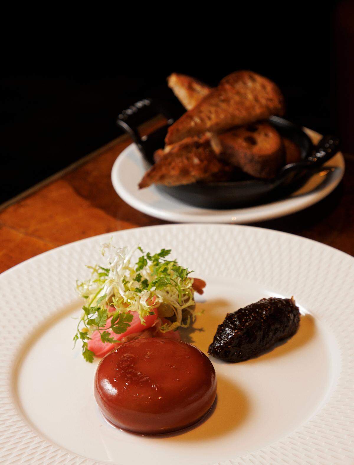 A close-up, angled shot of a gourmet appetizer on a white, textured plate. In the foreground, a round, glossy, reddish-brown pâté sits next to a small salad of frisée and a quenelle of dark jam. In the blurred background, a black dish filled with toasted crostini rests on a wooden table.