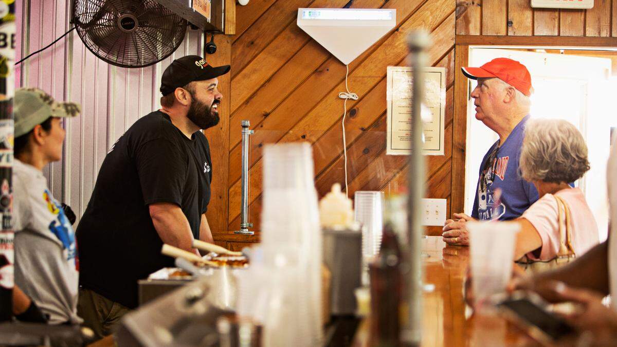 Firekeeper Jon Garren Kirkman, center, takes first-time customer Tom Cone’s order at Jon G’s Barbecue Saturday morning, June 25, 2022. The Peachland restaurant is only open on Saturdays and attracts a crowd from around the state.