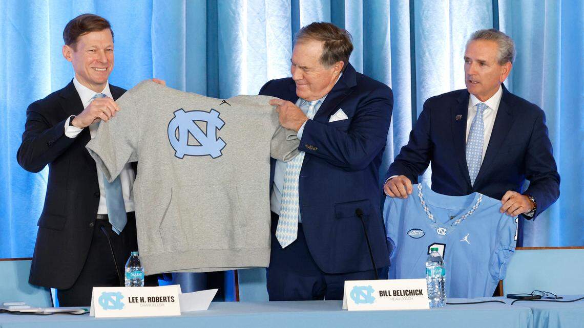 UNC Chancellor Lee Roberts gives new North Carolina head football coach Bill Belichick a hoodie with cut off sleeves during a press conference at the Loudermilk Center at UNC in Chapel Hill, N.C., Thursday, Dec. 12, 2024. UNC AD Bubba Cunningham stands to the right.