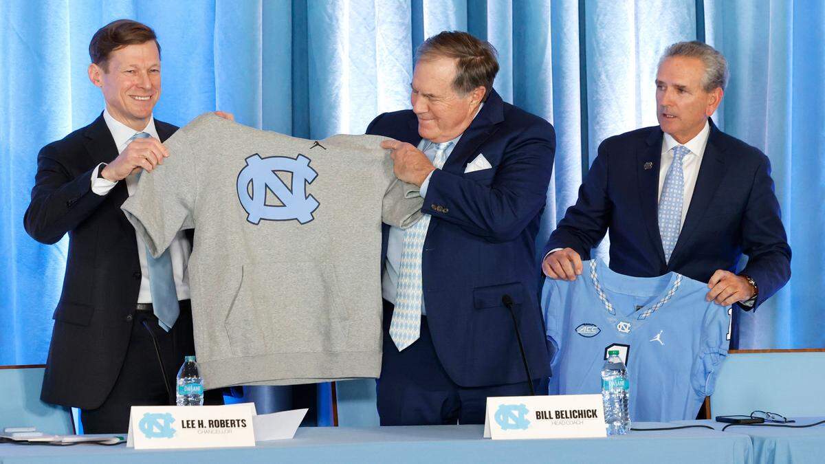 UNC Chancellor Lee Roberts gives new North Carolina head football coach Bill Belichick a hoodie with cut off sleeves during a press conference at the Loudermilk Center at UNC in Chapel Hill, N.C., Thursday, Dec. 12, 2024. UNC AD Bubba Cunningham stands to the right.