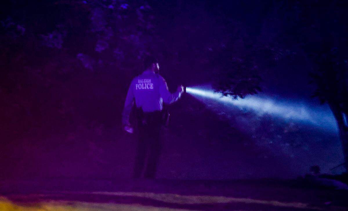 A Raleigh Police officer shines a light behind homes in the Hedingham neighborhood and Neuse River Trail area in Raleigh after 5 people were shot and killed Thursday, Oct. 13, 2022.