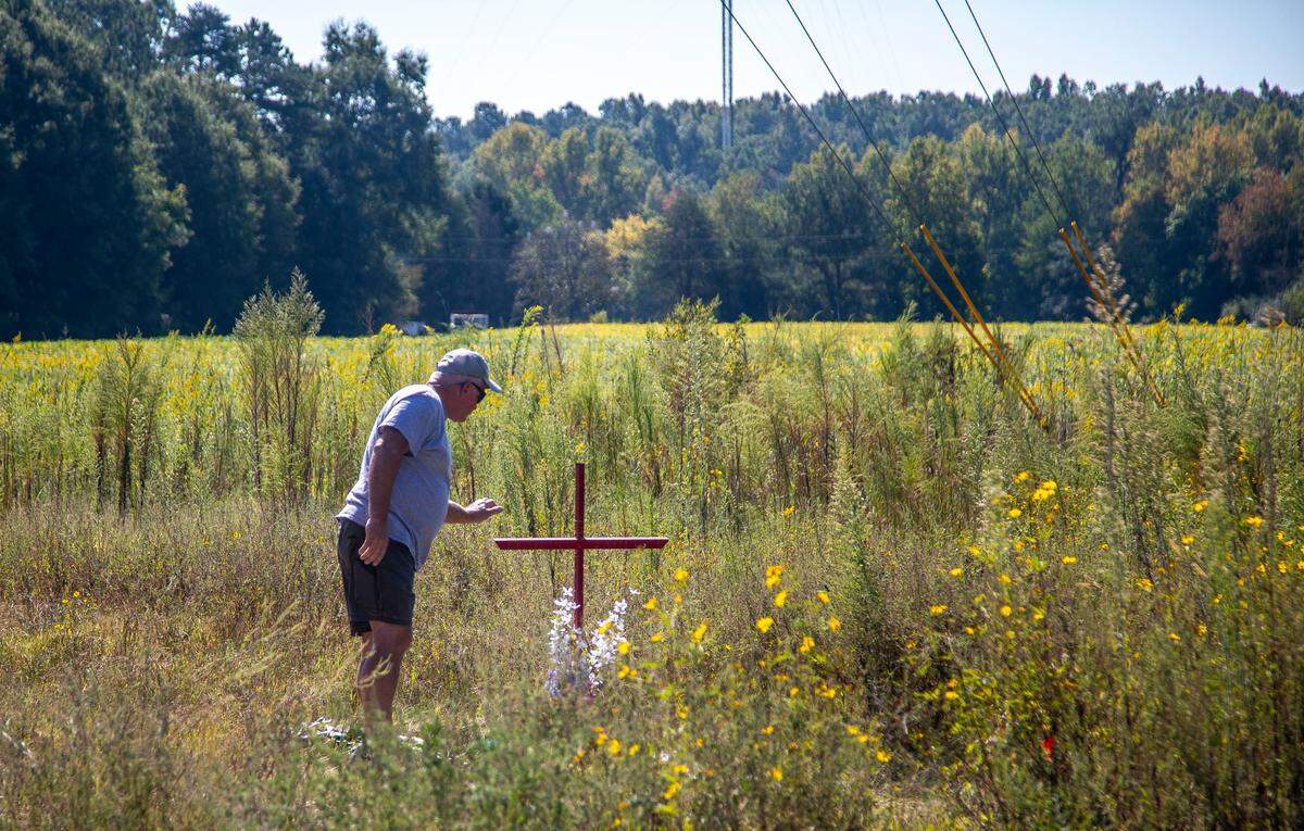 Stan Dean, the grandfather of Lyric Woods, reaches out to straighten a cross at a makeshift memorial that sits a few yards from the entrance of a gravel road in Orange County Tuesday, Sept. 20, 2022. The Orange County Sheriff’s Office said Monday the two young people found shot and killed Sunday in western Orange County are missing teens Lyric Woods, 14, and Devin Clark, 18.