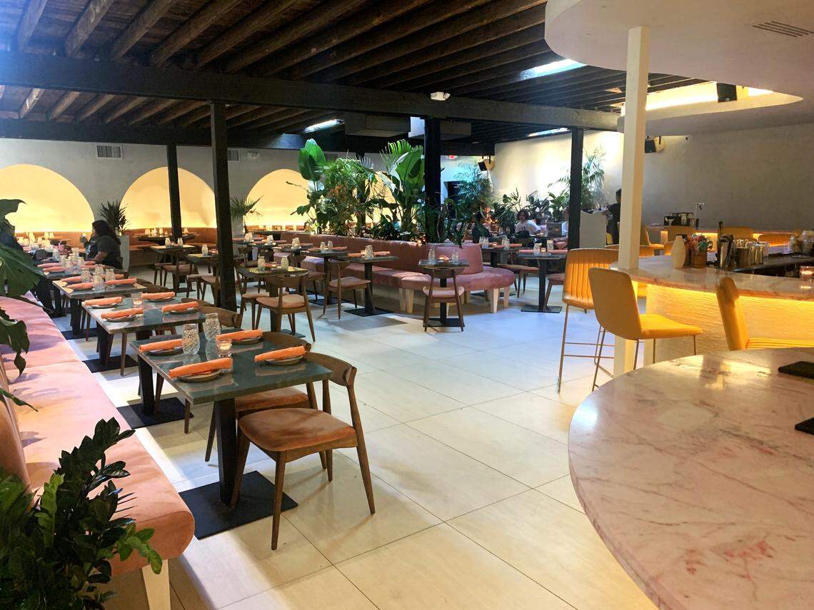 A sweeping view of a chic restaurant’s interior. The space is characterized by an open, dark wood-raftered ceiling and is filled with green plants. Pink upholstered banquettes line the walls, and a curved, warmly lit bar with a pink marble top and yellow stools is on the right.