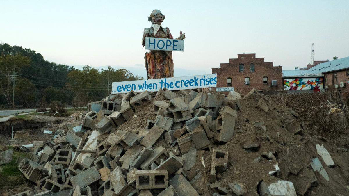Asheville’s River Arts District suffered some of the most catastrophic damage during Hurricane Helene. A city official said 80 percent of the district was destroyed. In the days after the storm a makeshift statue was erected off of Lyman Street, next to the French Broad River. The figure stood atop a pile of cinder blocks and held a sign that spoke not only for the city, but the entire region.