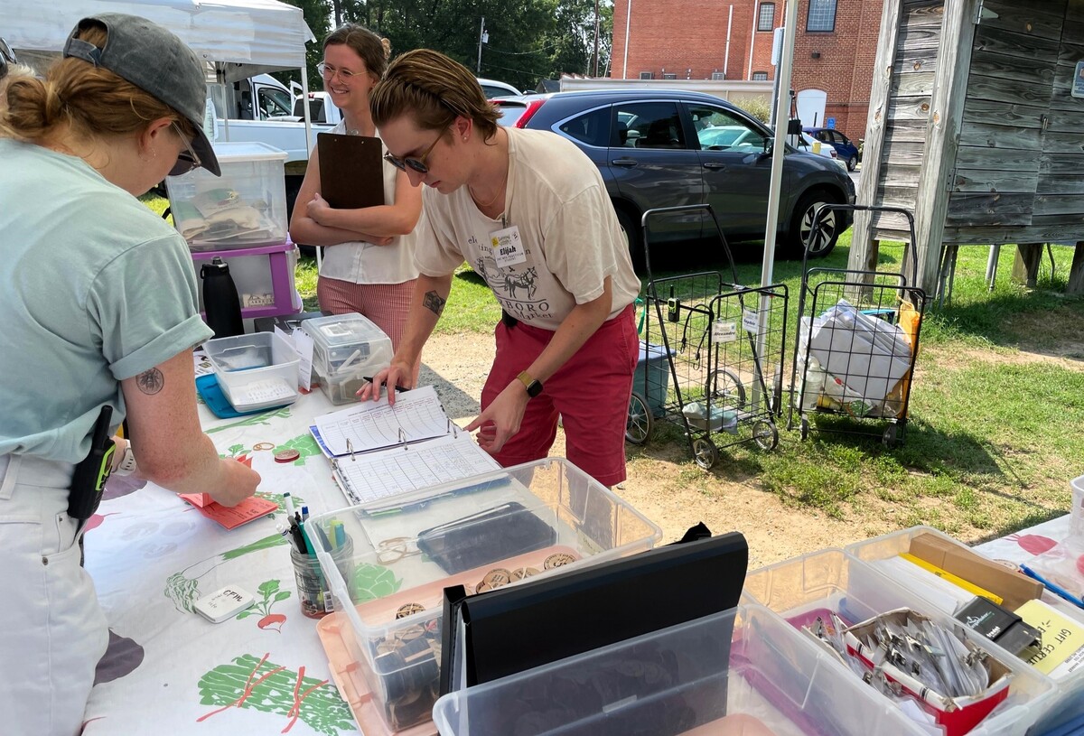 Elijah Moracco-Schelp (at right), a UNC-Chapel Hill public health student, and Carrboro Farmers’ Market manager Maggie Funkhouser help a customer with Seniors Farmers Market Nutrition Program food benefits get Double Bucks tokens on Wednesday, July 31, 2024.