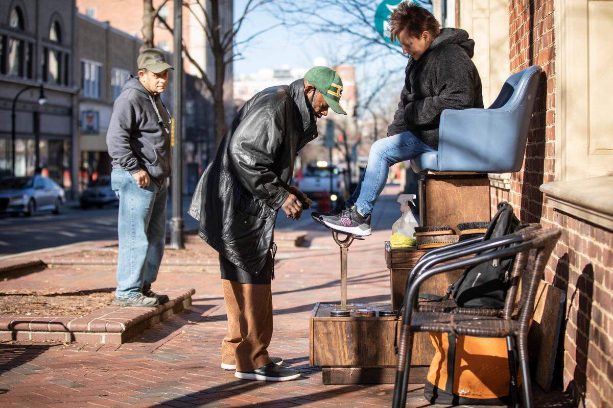 Pierce McKoy, a Durham shoe shiner, works on brightening the soles of customer Cathy Brown’s shoes in his newly refurbished chair on Main Street in Durham, N.C. while her husband Kent Brown, left, watches on Monday, Dec. 13, 2021.