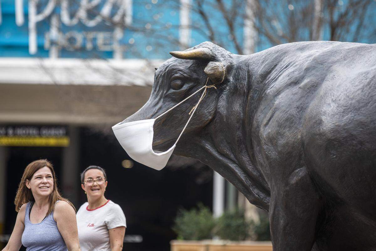 Pedestrians pass the “Major” the Durham bull bronze sculpture now sporting a protective mask in the CCB Plaza on Thursday, Mar. 19, 2020, in Durham, N.C.