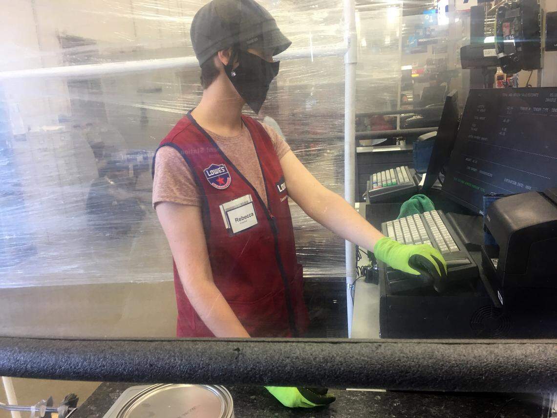 An employee wearing a mask and gloves rings up a sale behind a plastic shield at the Lowe’s in Garner.