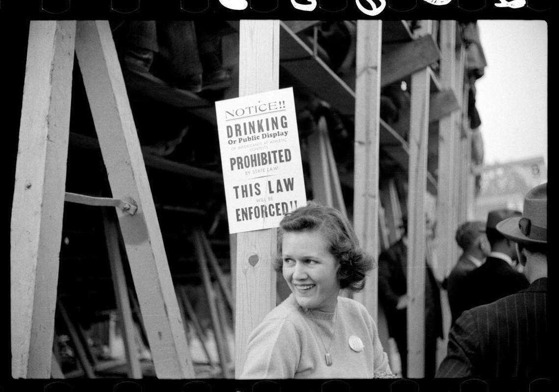 A UNC fan wearing a “Beat Duke” button stands beneath a sign prohibiting drinking alcohol under the bleachers at the Duke-Carolina game played in Durham, NC in 1939.