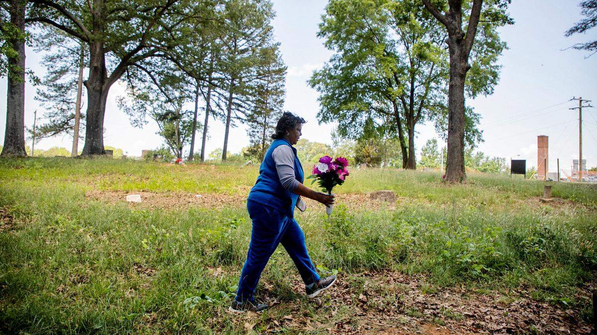 Deborah Lofton carries over a new bouquet of plastic flowers to mark the unmarked graves of her great grandparents under a tree in the Oak Grove Cemetery, which was founded by freed African-Americans after the Civil War and now has lost more of its tree buffer to widening of the I-440 Beltline, that separated it from the rest of the Method community in the 1960s, on Saturday, May 8, 2021, in Raleigh, N.C.