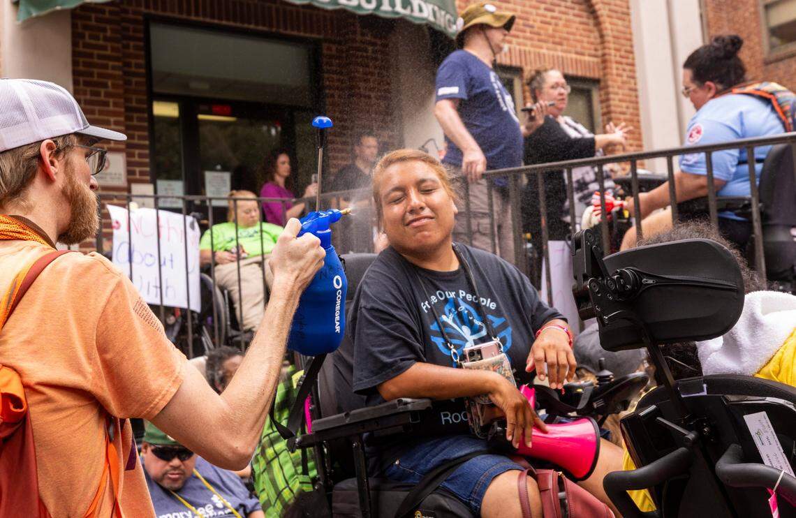 Joe Tate, a personal care assistant, mists Lucero Torres, among demonstrators rallying outside the North Carolina Department of Health and Human Services on Monday, June 24, 2024. About 50 demonstrators with the National Disability Rights Organization ADAPT rallied outside the office to mark the 25th Anniversary Rally of the Olmstead Decision in North Carolina. The group aims to raise awareness about the decision’s importance and advocate for shifting investment from facility-based to community-based services in North Carolina, “To assure the civil and human rights of people with disabilities to live in freedom,” according to Disability Rights North Carolina’s website.