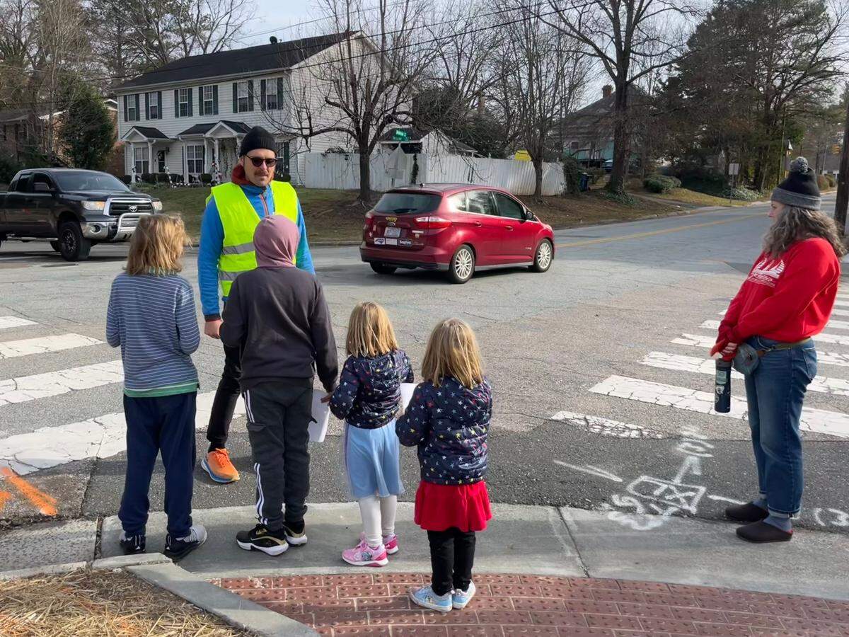 Jacopo Montobbio, in yellow, instructs students to wait for cars at an intersection near Lakewood Elementary School on Saturday, Dec. 14, 2024, in Durham, North Carolina. Crossing guards can be identified by their official uniforms.