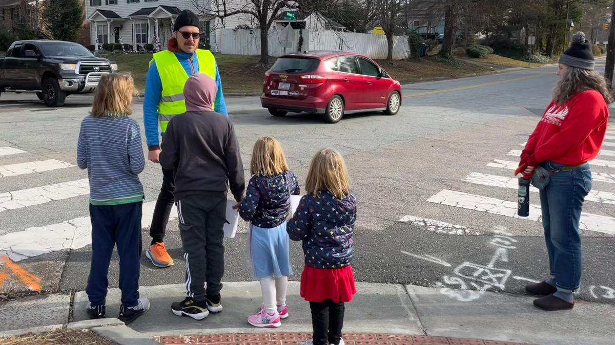 Jacopo Montobbio, in yellow, instructs students to wait for cars at an intersection near Lakewood Elementary School on Saturday, Dec. 14, 2024, in Durham, N.C.