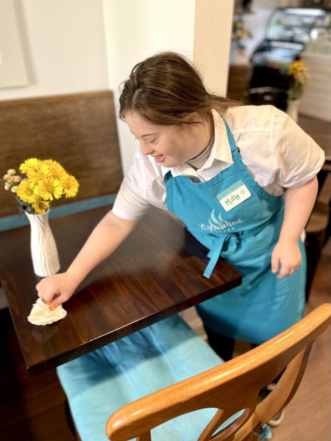 A young woman named Molly, wearing a bright blue “Esteamed Coffee” apron, smiles warmly while wiping down a dark wood café table. In the background, a white vase filled with cheerful yellow flowers sits on the table, adding to the bright and welcoming atmosphere.