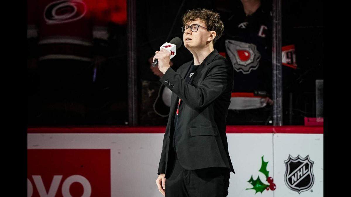 Mason Greer, wearing a black suit jacket with a Carolina Hurricanes jersey underneath, holds a Canes-branded microphone while he sings the national anthem.