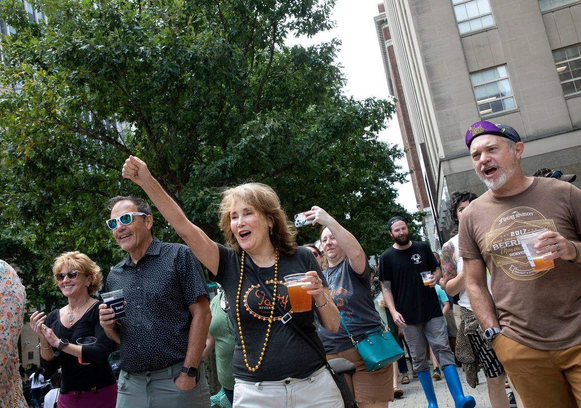 Susan Carl raises a fist in applause as a second line band leads a group down Fayetteville Street from Crank Arm Brewing Company to The Pour House Music Hall and Record Shop on the first day of Raleigh’s social district on Monday, Aug. 15, 2022. Participating businesses in the district that serve alcohol will be able to sell alcoholic beverages that people can drink as they walk to their next location.