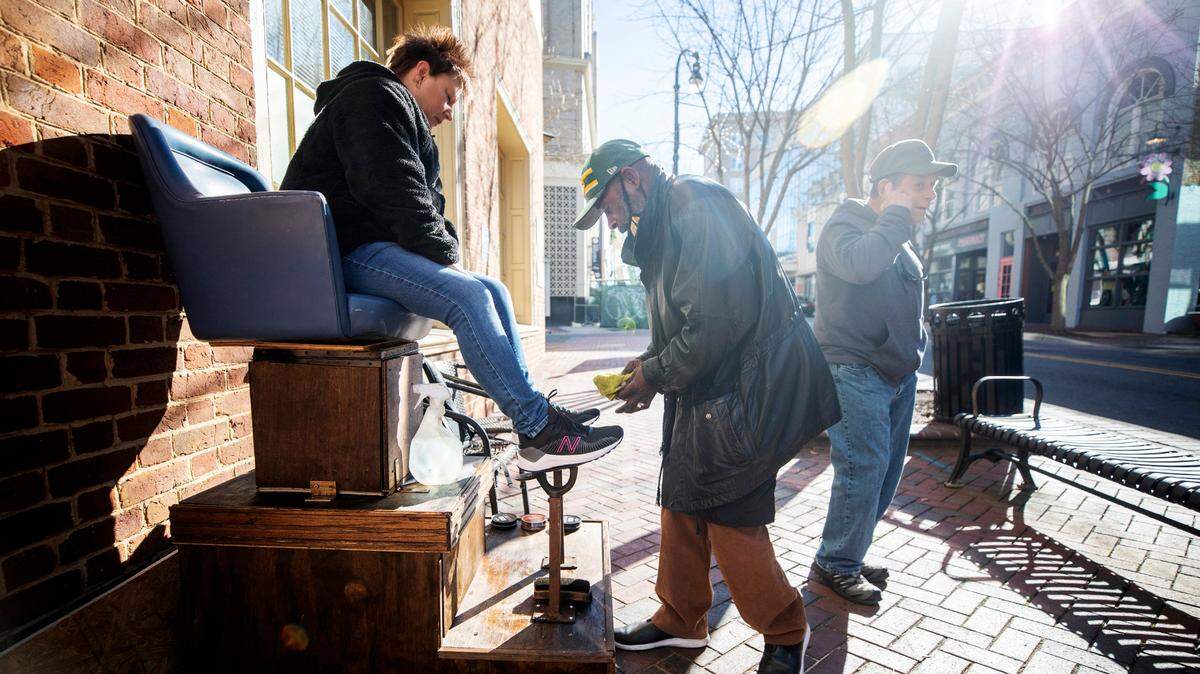 Cathy Brown sits in Durham shoe shiner Pierce McKoy’s newly refurbished chair on Main Street in downtown Durham, N.C. while her husband Kent, right, waits for her on Monday, Dec. 13, 2021. Local residents recently raised roughly $2,500 to help McKoy get the chair refurbished.