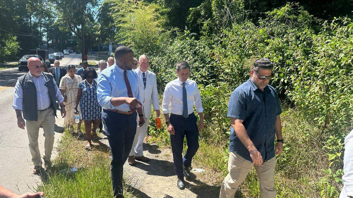 Durham Mayor Leonardo Williams and U.S. Transportation Secretary Pete Buttigieg walk on a gravel path along Holloway Street on Tuesday, July 2, 2024. The city will build sidewalks, crosswalks and new bus shelters along 2 miles of Holloway using a $12 million federal grant Buttigieg announced a few days earlier.