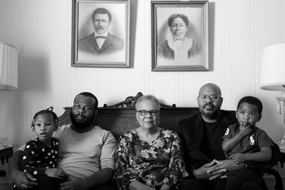 Toney and Nellie Strayhorn, both freed from slavery, purchased a small plot of land on the outskirts of Chapel Hill in Carrboro, NC, named after white supremacist Julian Carr, in the mid 1870Õs. They built a one-room cabin that eventually expanded to a two-story home. The family survived and thrived through the racial violent times of Reconstruction and Jim Crow. Dolores Clark (centered) sits with her children and great-grandchildren in the home her great-grandparents, Toney and Nellie Strayhorn (pictured on the wall), built around 1879 in Carrboro, North Carolina.