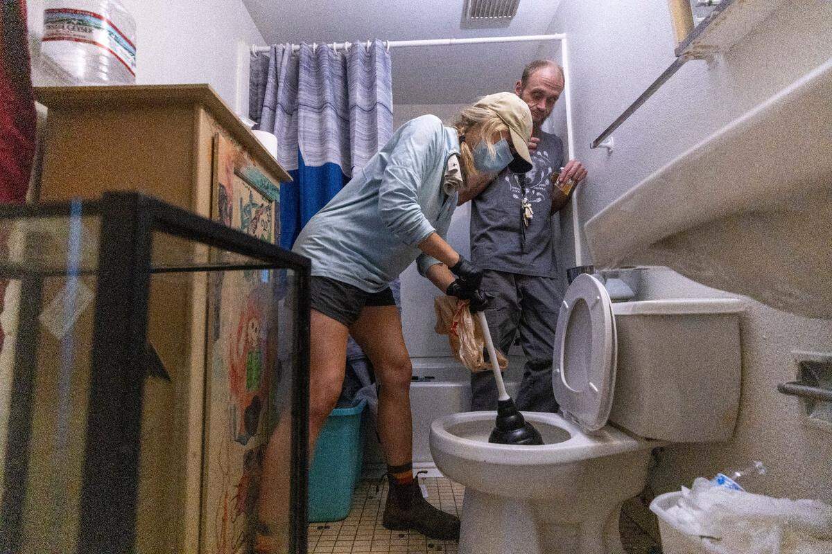 Hannah Huston, a volunteer with grassroots group BeLoved Asheville, unclogs a toilet for Jesse Cook who lives in a iow-income independent living facility, with no running water on Wednesday, October. 10, 2024. About half of Asheville’s water customers were still without running water this week because the remnants of Hurricane Helene ripped apart the city’s distribution system. Volunteers haul gray water in 5-gallon buckets to flush toilets to help reduce the risk of illness.
