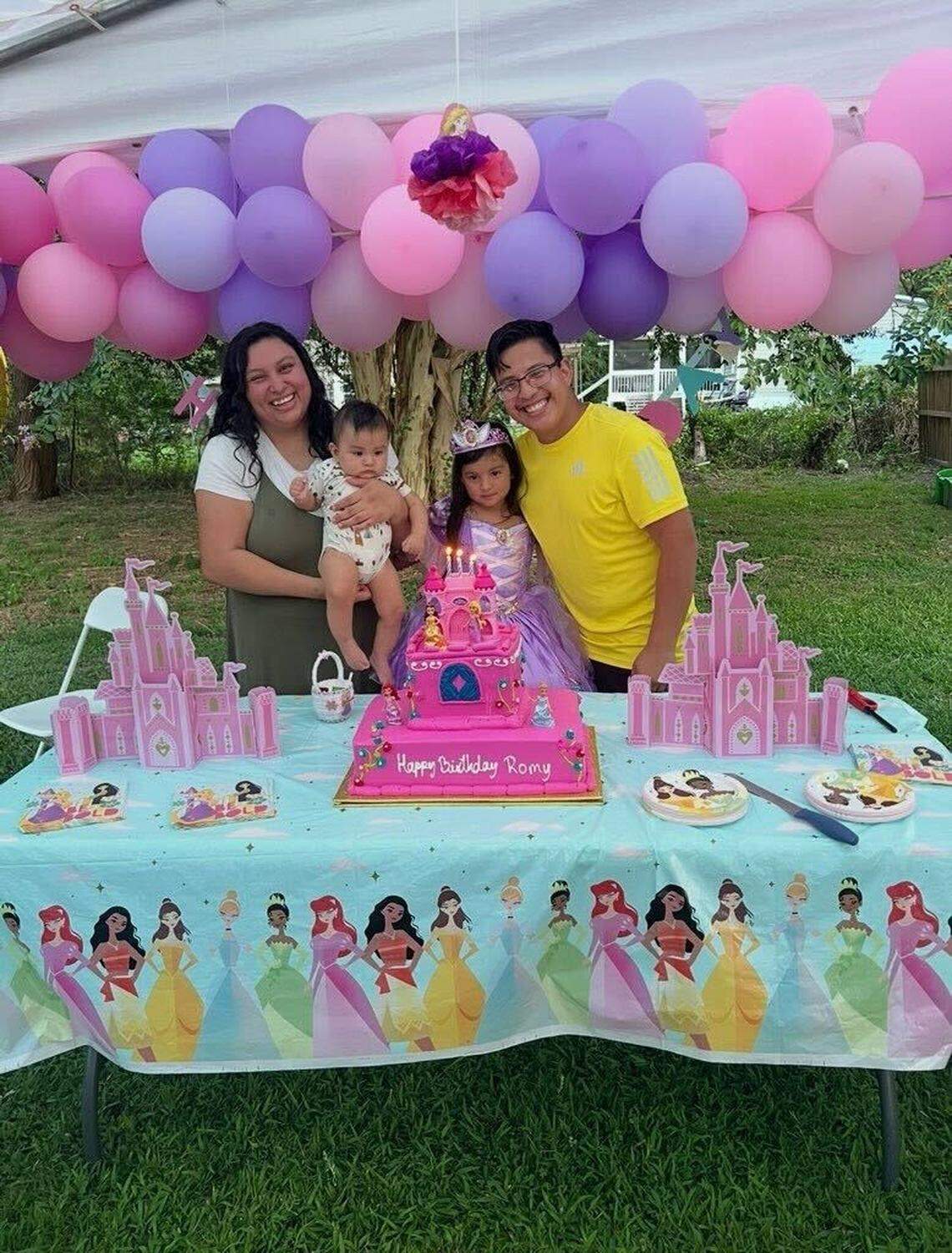 Moises Benitez Diaz, in a yellow shirt and glasses, embraces his daughter, Rochelle in one arm as they pose for a picture at Rochelle’s birthday party. His wife, Esmeralda Escobar, in green overalls, is holding their son, Elijah. The party is Disney princess-themed, with a decorated blue table cover featuring Disney princesses like Tiana, Moana, and Cinderella. Rochelle wears a lavender tiara and gown behind a pink cake that reads “Happy birthday Romy!”