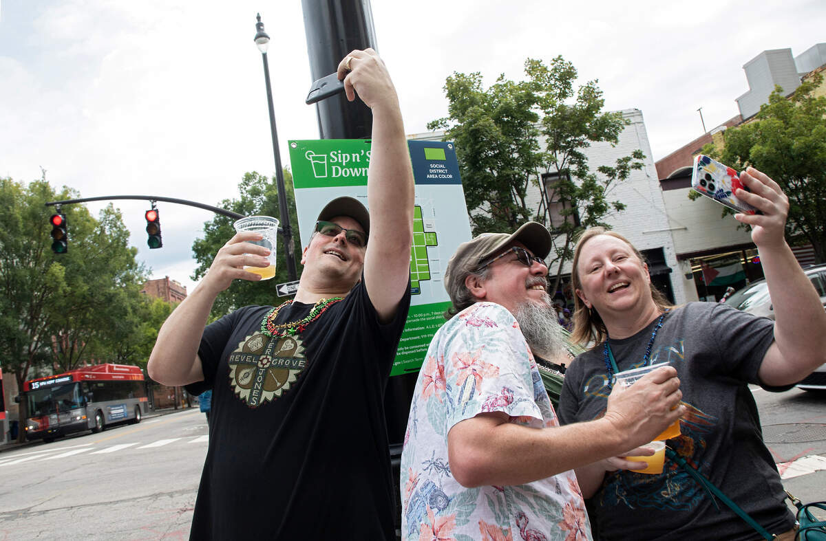From left, Rob Moffett, Ryan Kempf and Valerie Kempf take selfies on the first day of Raleigh’s social district on August 15, 2022.