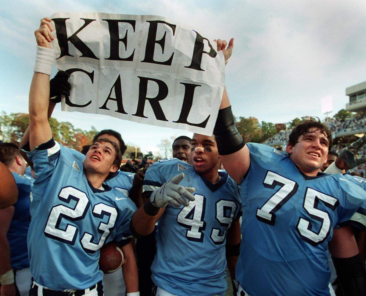 UNC players hold up a “Keep Carl” sign during their victory celebration after the Tar Heels defeated Duke 38-0 in Kenan Stadium in 1999. The players borrowed the sign, which refers to embattled head coach Carl Torbush, from a fan in the endzone seats. From left: Brian Schmitz (23), Julius Peppers (49) and Allen Mogridge (75).