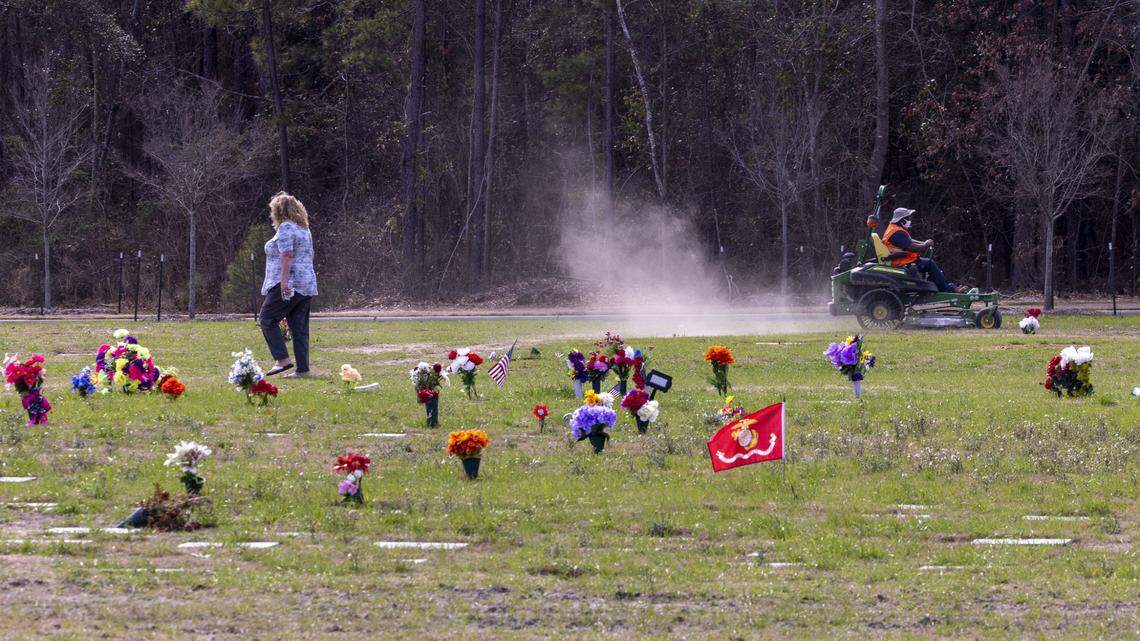 A mourner visits a gravesite as a groundskeeper mows grass at the Sandhills State Veterans Cemetery in Spring Lake Tuesday, Feb 21, 2022. North Carolina’s four state-owned veterans cemeteries are behind on maintenance and are short-staffed.