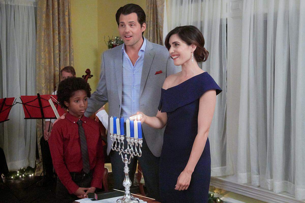 Career-minded Rebecca’s plans for Hanukkah go askew when a promotion opportunity comes up at work. When the company CEO asks Rebecca and her insufferable office mate, Chris – also her main competitor for the promotion – to plan the company’s Christmas party, she realizes they must overcome their opposing styles in order to succeed. Forced to work together on the holiday party, Chris learns more about Rebecca and her family’s Hanukkah traditions, while she also begins to see him in a new light. Although feelings slowly develop between the two, the ongoing competition over the promotion threatens to undermine it all. Photo: Ceyon Crossfield, Kristoffer Polaha, Carly Pope Credit: ©2019 Crown Media United States LLC/Photographer: Albert Camicioli