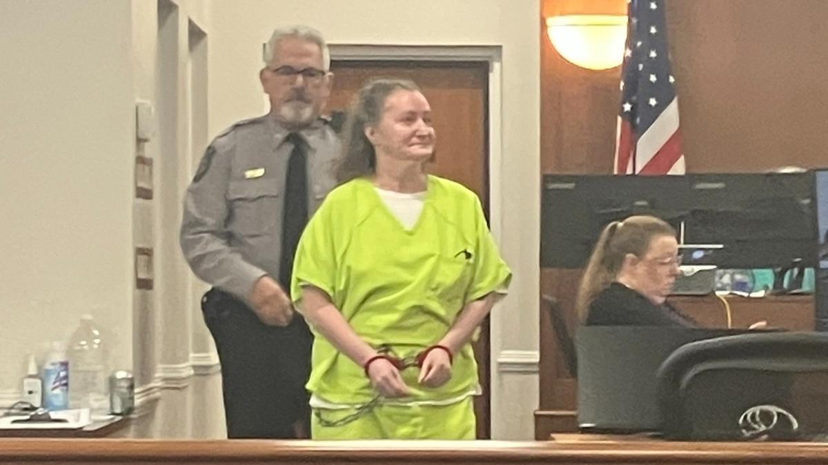 Paula Decoteau, a middle-aged woman with a ponytail, is in front of a bailiff in a gray shirt and black tie. She wears a bright lime green jumpsuit and bearing chains around her wrists and midriff as she sits in the Battle Courtroom of the Orange County Courthouse.
