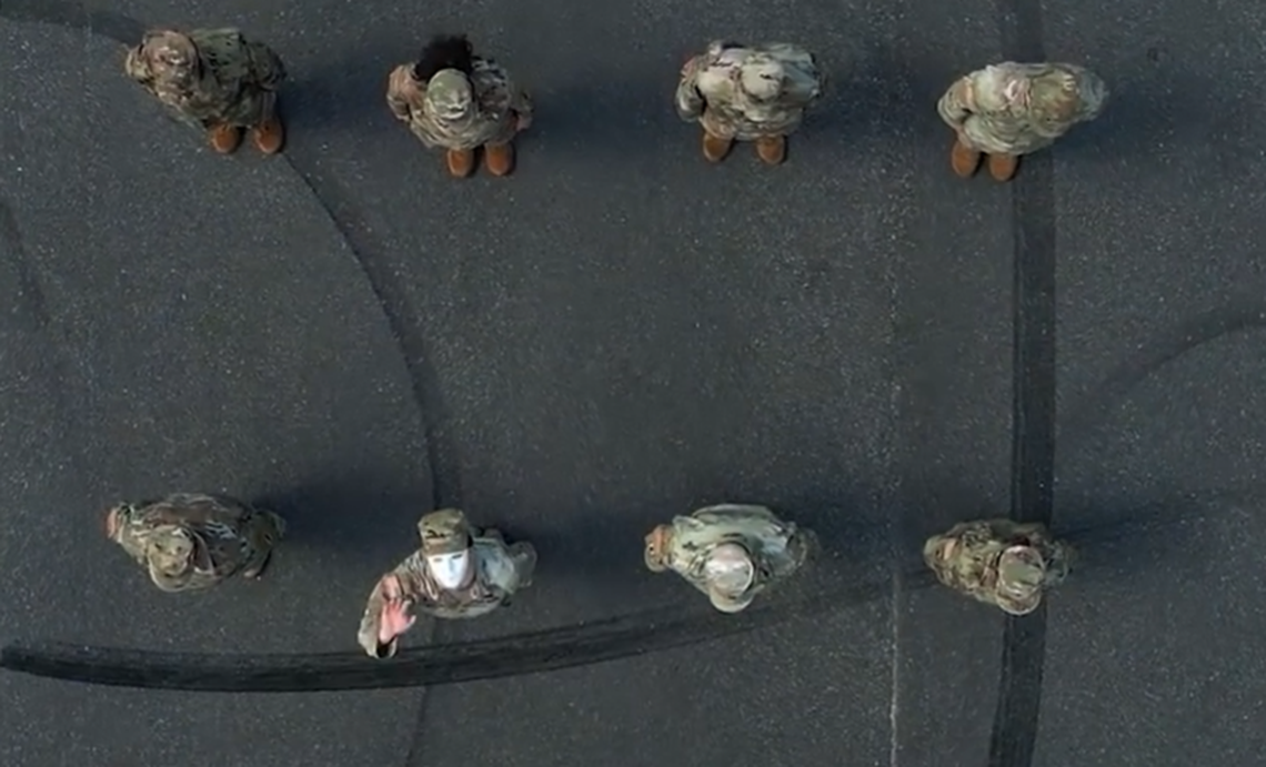 In this image from the video, a masked figure is seen waving at a drone hovering over head.